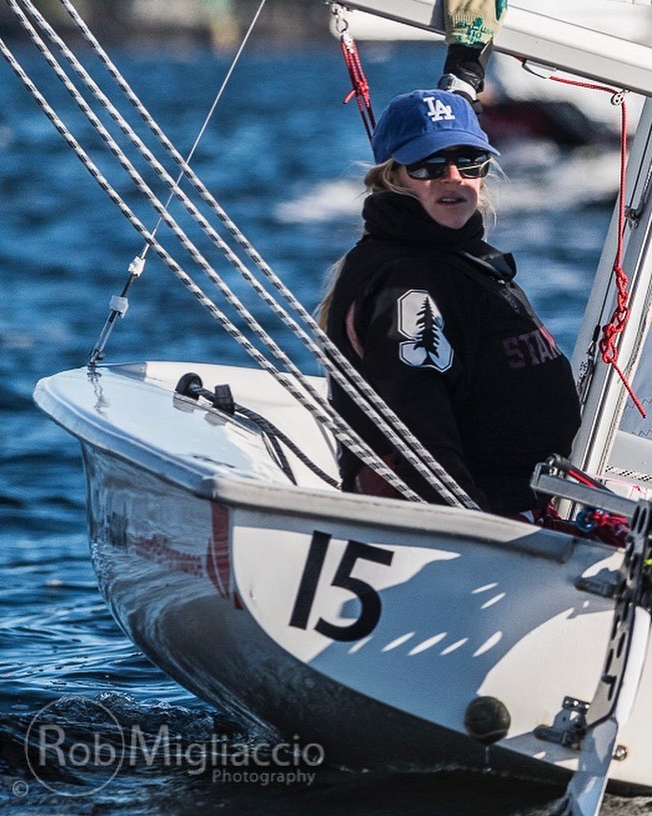 A person wearing sunglasses, a blue LA baseball cap, and a black Stanford jacket is sailing a small white boat marked number 15 on a body of water. Sail ropes and waves are visible. The image is credited to Rob Migliaccio.