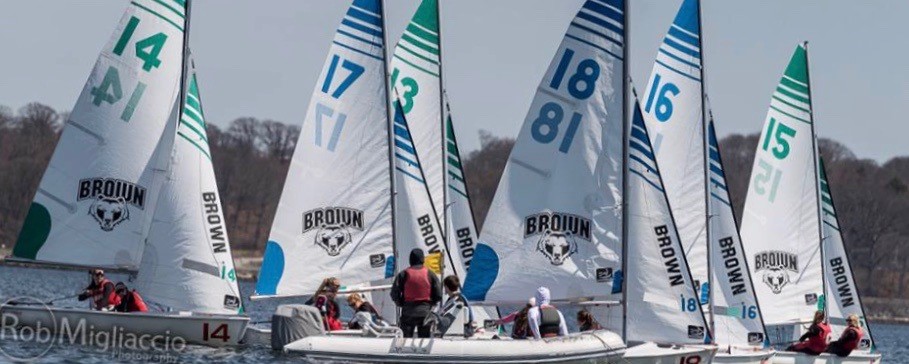 A group of sailboats with 'Brown' logos and numbered sails compete on a calm body of water, each boat crewed by two people under a partly cloudy sky. Trees are visible in the background.