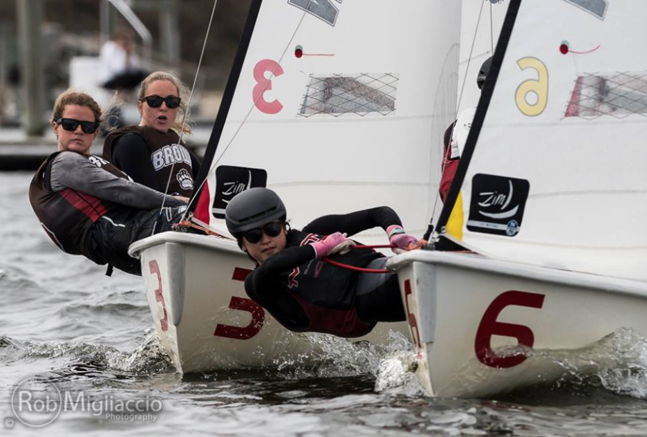 Two teams of sailors compete in small dinghies, leaning out over the water to balance their boats. The sailors wear helmets, life jackets, and team gear. The boats are marked with the numbers 3 and 6.