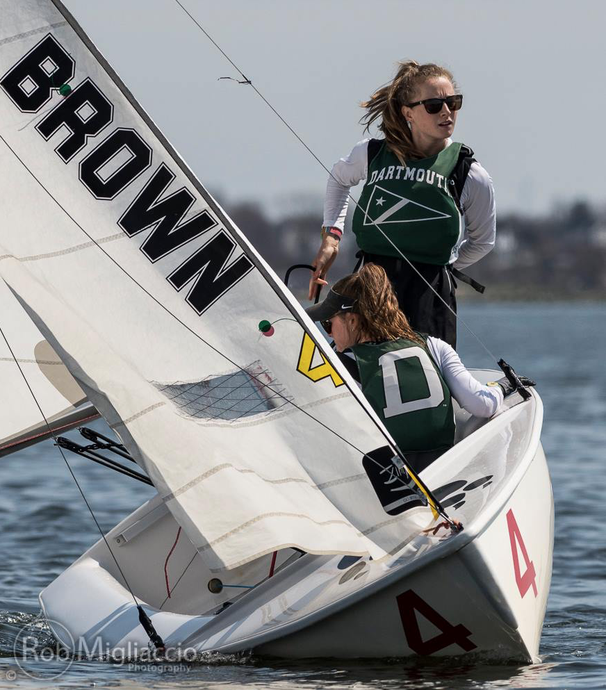 Two female sailors in Dartmouth gear steer a small sailboat with a sail labeled "BROWN" and the number 4. The boat is tilted on water, and both sailors appear focused on the competition.
