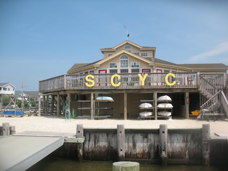 A tan, two-story waterside building with large yellow letters 'SCYC' on the balcony railing. Several kayaks and rowboats are stored underneath, with docks and calm water in the foreground.