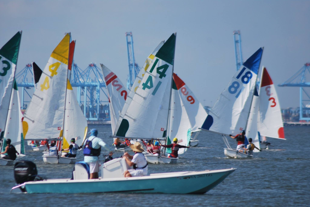 Numerous small sailboats with colorful numbered sails race on the water, while people in a motorboat observe. Industrial cranes are visible in the background against a blue sky.
