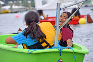 Two children wearing life jackets sit in a small green boat on the water, smiling and laughing together. Other boats and blurred figures are visible in the background.