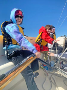 Three children wearing life jackets and colorful jackets sit on a sailboat, smiling and steering the boat on a sunny day with a clear blue sky. One child holds the tiller, while others hold the ropes.