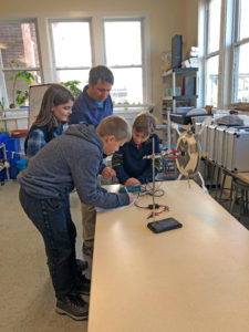 Three kids and an adult stand around a table in a classroom, working together on a science experiment involving a small wind turbine and electronic equipment. The room is bright with large windows in the background.