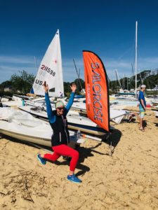 A person in bright athletic clothing kneels on sandy ground with arms raised, smiling near a "SAILORSJC" flag and sailboats, under a clear blue sky. Another person stands nearby among the boats.