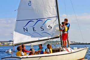 Six people wearing life jackets sail a small boat on a sunny day. One person adjusts the sail labeled "NESS" while others sit or steer. The water is calm, and buildings and trees are visible in the background.