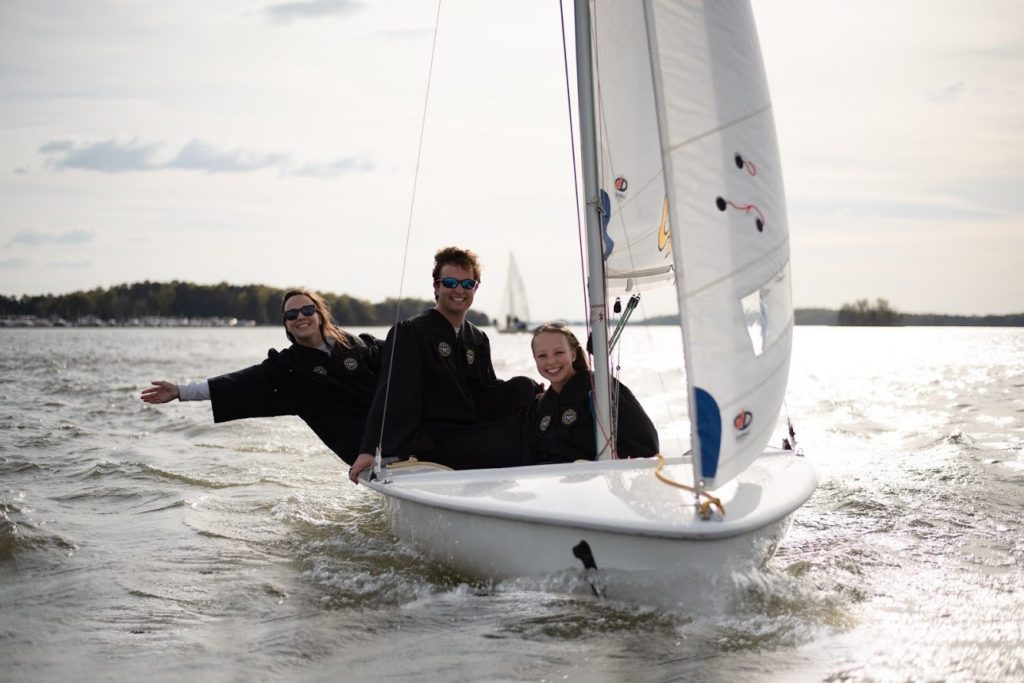 Three people in jackets smile and pose on a small sailboat on a lake, with one person leaning out over the water. The sun is bright, and distant trees and another sailboat appear in the background.