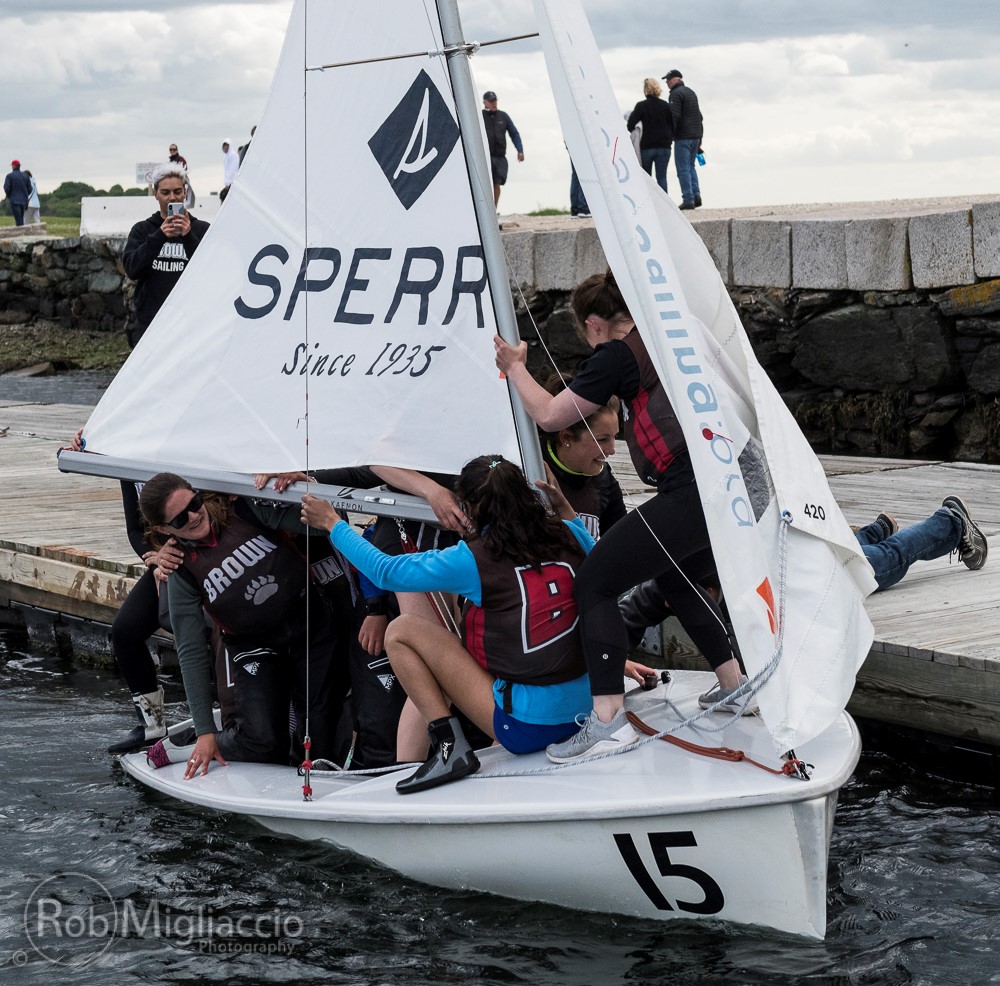A group of young sailors in life jackets crowded on a small sailboat labeled '15' and 'SPERRY' maneuver near a dock, with some holding the sail and one pushing from the dock.