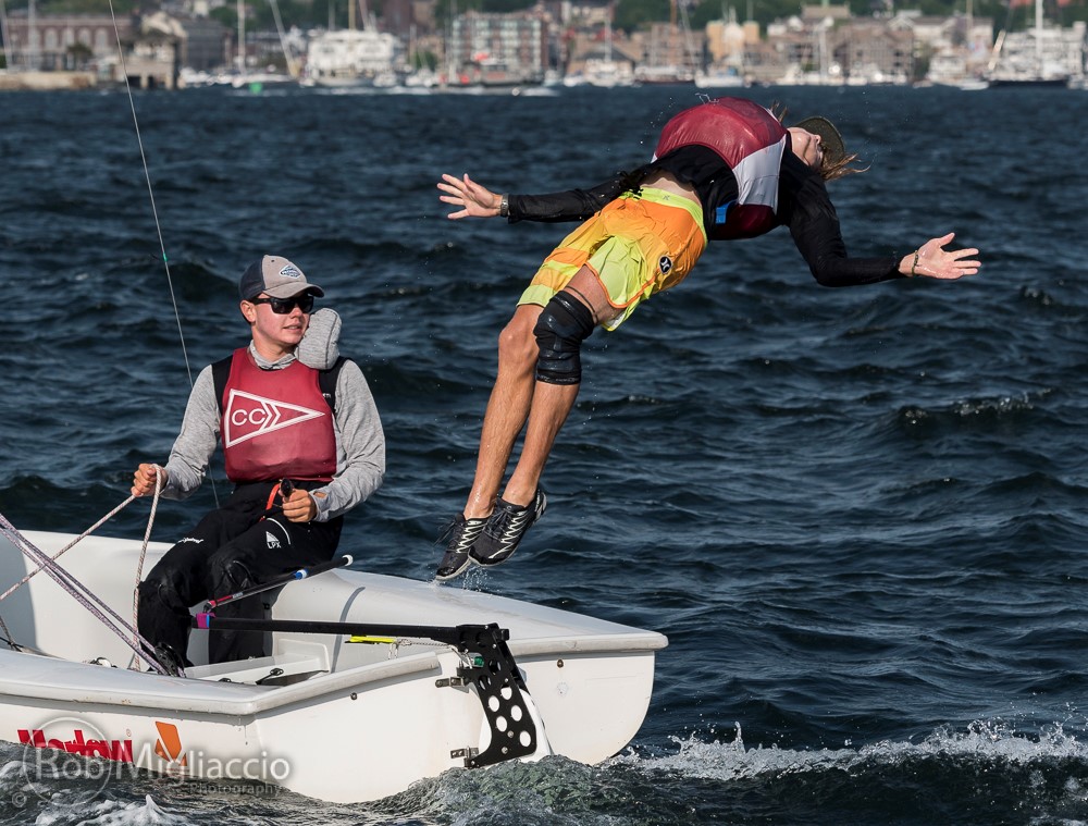 A man in colorful shorts falls backward off a small sailboat into choppy water, while another person wearing a cap and sunglasses sits in the boat, watching. Buildings and trees are visible in the background.