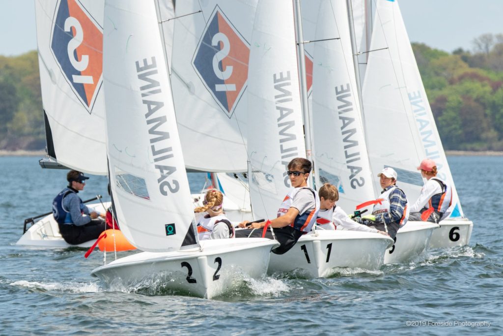 A group of young sailors compete in small sailboats on calm water, each boat marked with numbers and “SAILMAINE.” Trees are visible in the background under clear skies.