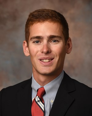 A young man with short brown hair, wearing a dark suit, light blue shirt, and red striped tie, smiles in a professional headshot with a neutral brown and gray background.