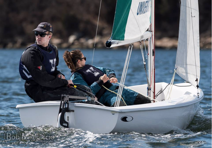 Two people in life jackets sail a small white dinghy on a sunny day, with water and blurred trees in the background. One person steers while the other adjusts a rope.