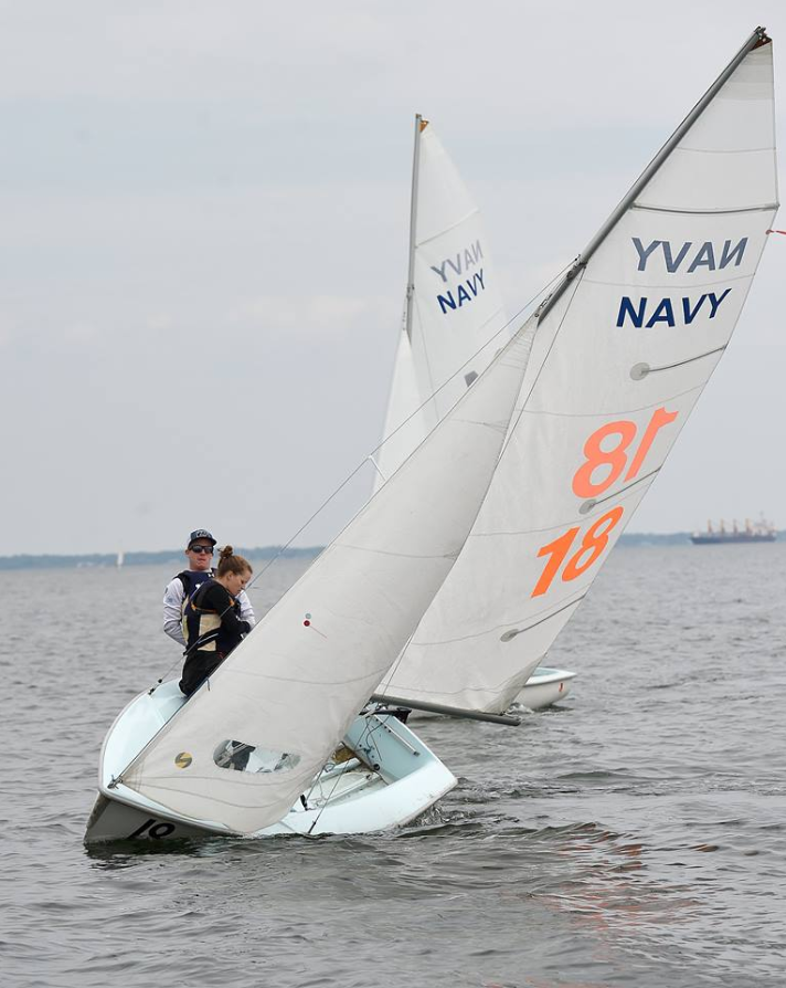 Two small sailboats with white sails marked "NAVY 18" glide on calm water. Three people are visible in the foreground boat, wearing life jackets, with another sailboat in the background. The sky is overcast.