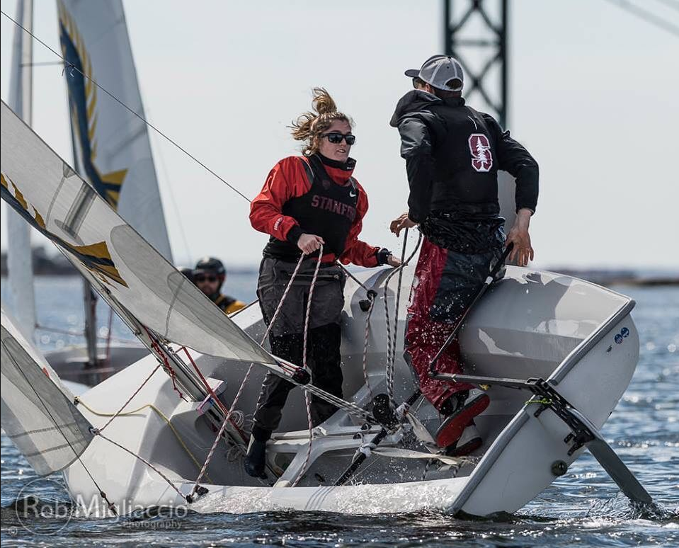 Two sailors in Stanford gear balance on the edge of a tilting sailboat, gripping ropes as water splashes on deck during a sailing race. Another sailor and part of the mast are visible in the background.