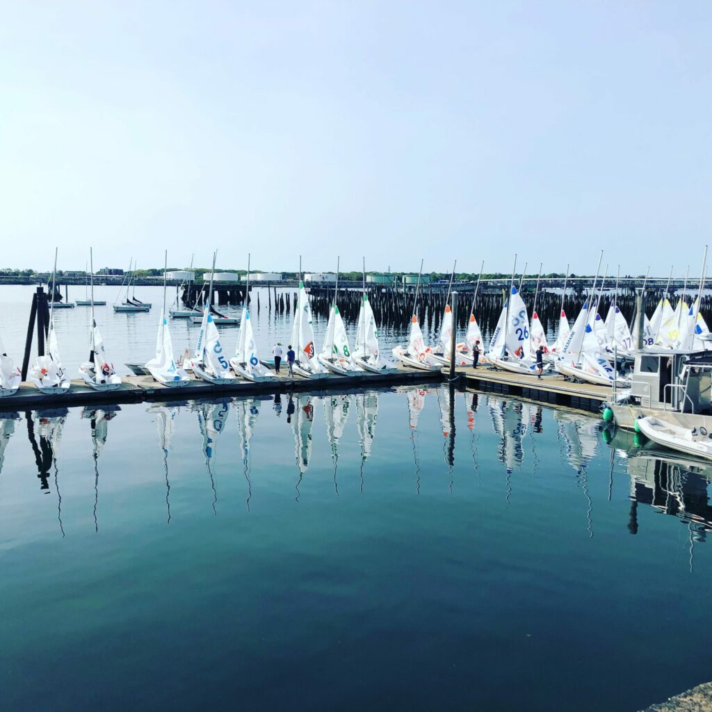 A row of small sailboats is docked at a marina on calm water, with their reflections clearly visible. People are gathered near the boats, and a hazy sky stretches above the distant shoreline and pier.