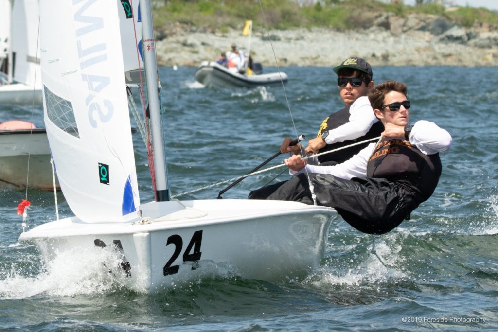 Two sailors wearing sunglasses and life jackets are leaning out of a small sailboat numbered 24, racing on choppy water with other boats and a rocky shoreline visible in the background.