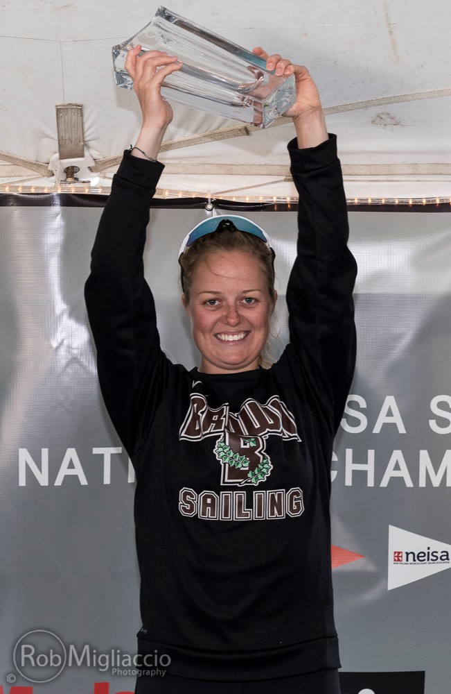 A smiling woman in a 'Bears Sailing' sweatshirt holds a clear trophy above her head, standing in front of a banner at a sailing championship event.