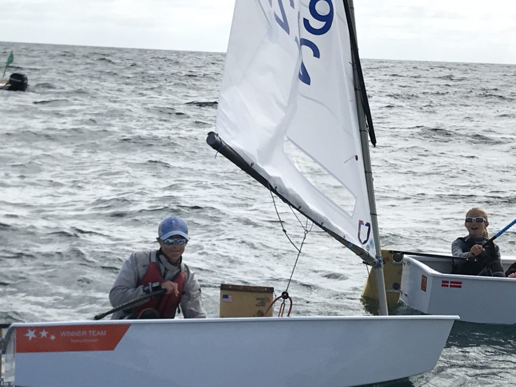Two people are sailing small dinghies on the open water. Both wear sunglasses and life jackets. The boat in the foreground has a partially visible sail and the words “WINNER TEAM” on the side.