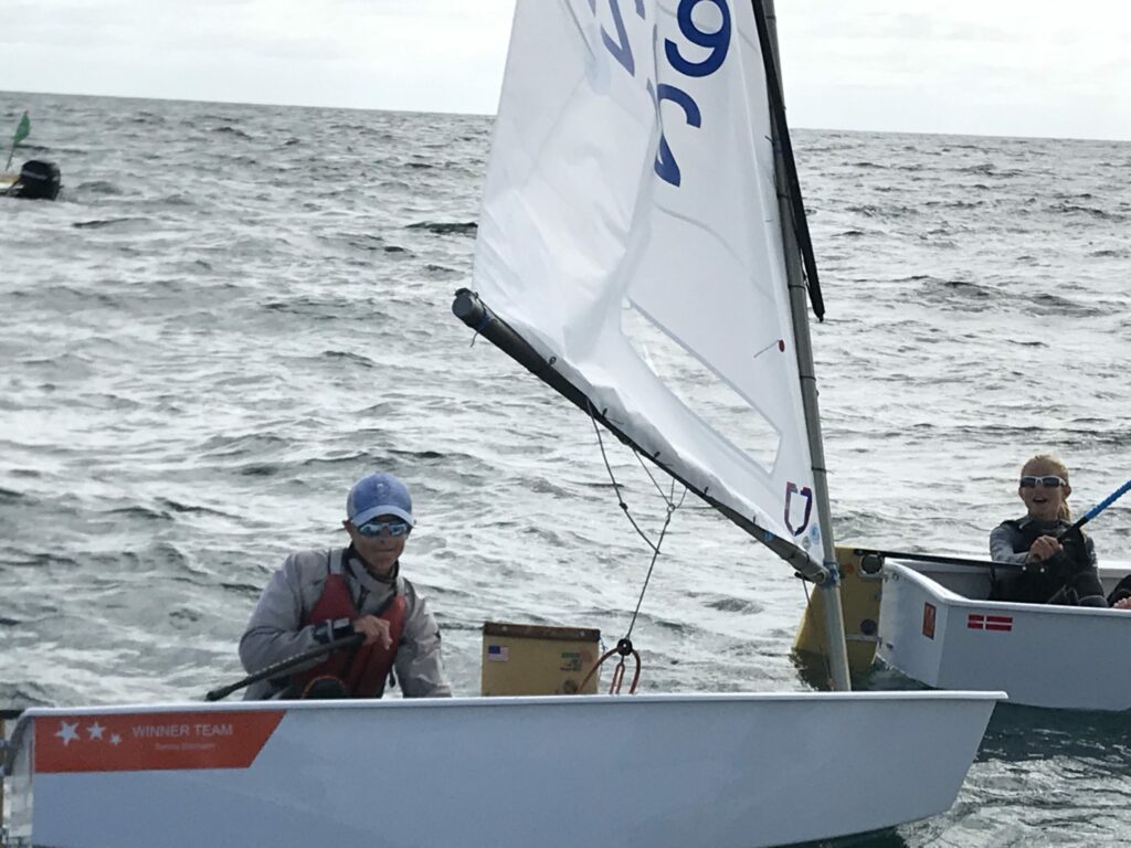 Two people are sailing small dinghies on the open water. Both wear sunglasses and life jackets. The boat in the foreground has a partially visible sail and the words “WINNER TEAM” on the side.