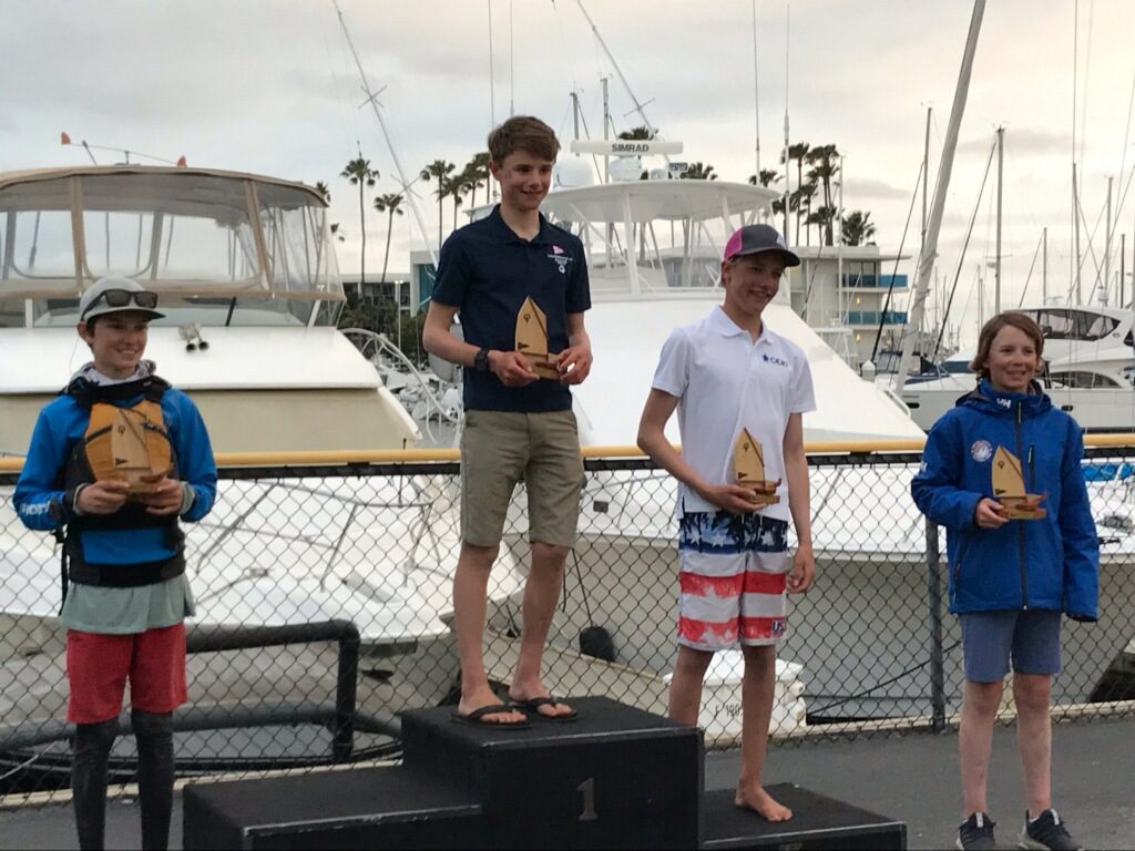 Four smiling teenagers holding trophies stand on a podium at a marina. Boats and palm trees are visible in the background. The teens are dressed in casual and sailing attire. The person in the middle stands on the tallest platform.