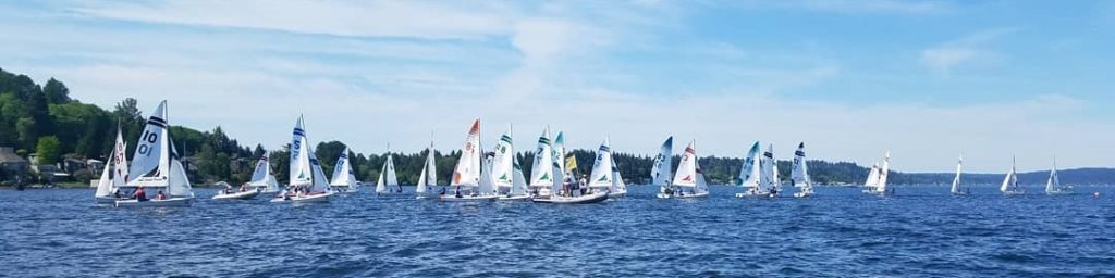 A group of small sailboats with colorful sails race on a blue lake under a partly cloudy sky, with green, tree-lined shores and houses visible in the background.