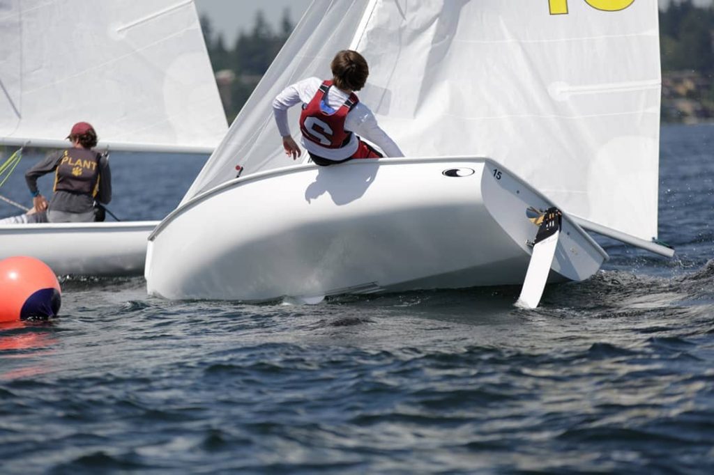 A sailor in a red life vest is leaning off the side of a capsizing sailboat on a lake during a race, with another sailor in a nearby boat and an orange buoy visible in the water.