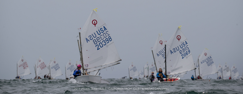 Youth sailors in small dinghies race on open water under cloudy skies, each boat displaying sail numbers, during the 2019 USODA Team Trials at California Yacht Club.