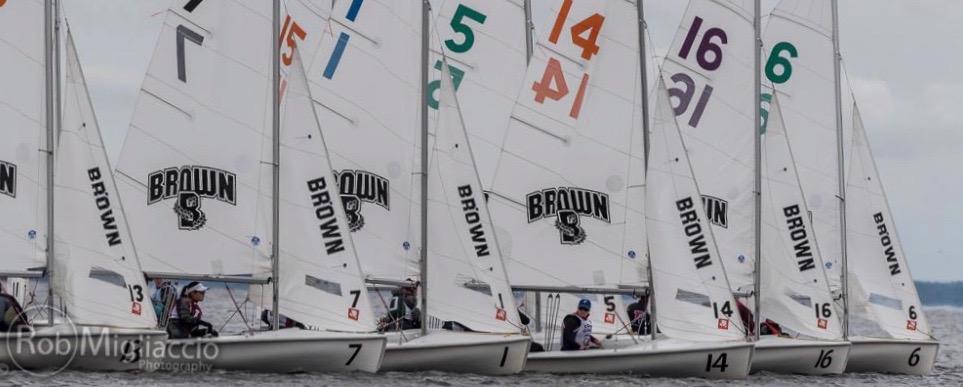 A close-up of several Brown University sailboats lined up side by side on the water, each with sail numbers and crews preparing to race on a cloudy day.