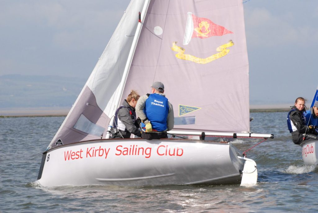 Two people sail a small boat with "West Kirby Sailing Club" written on the side, while another boat follows behind them on the water under a partly cloudy sky.