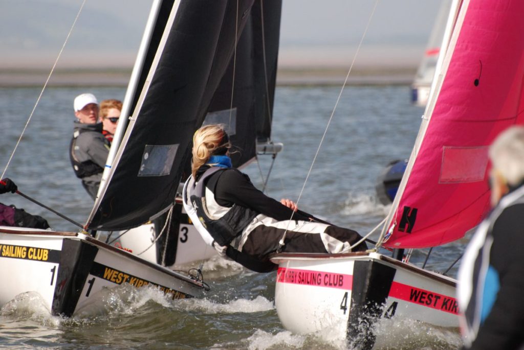 Three people are sailing small boats in a race on the water, wearing life jackets and harnesses, with sails labeled “West Kirby Sailing Club.” One sail is black and another is bright pink.