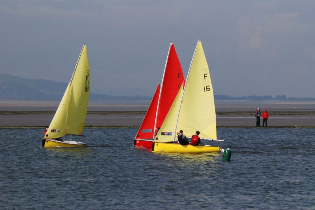 Three small sailboats, two yellow and one red, race on a body of water near the shore. Two people stand on the shore in the background, and hills can be seen in the distance under a cloudy sky.