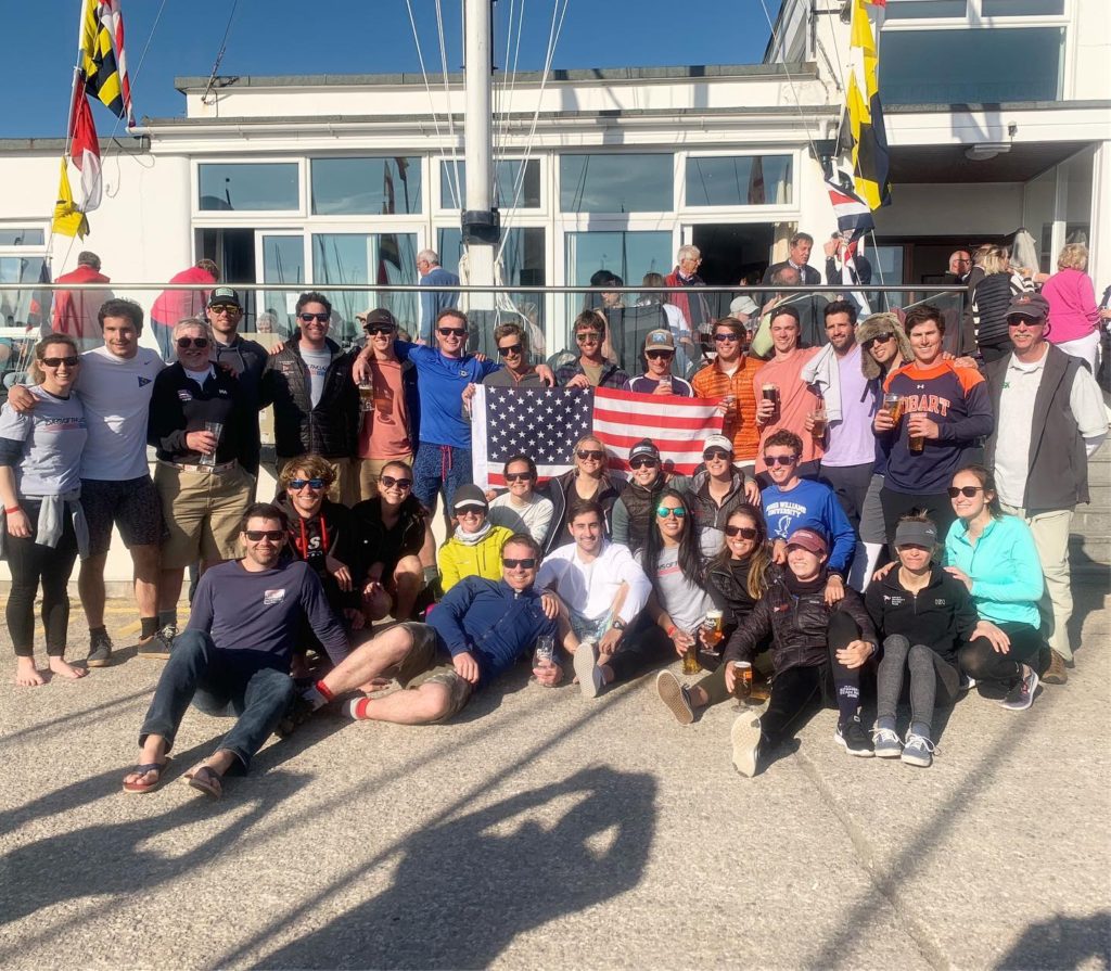 A large group of people pose together outside a building on a sunny day, holding two American flags and wearing casual, sporty clothing and sunglasses. Some are sitting, others standing, smiling at the camera.