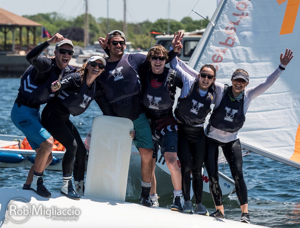Six smiling people in sailing gear stand together on a capsized sailboat, arms around each other, celebrating on the water under a sunny sky. A dock and other boats are visible in the background.