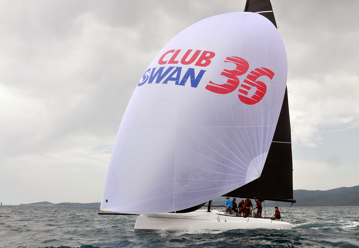 A white sailboat with a large sail displaying "CLUB SWAN 36" in red and blue letters glides on the ocean, with several people on board and a cloudy sky overhead.