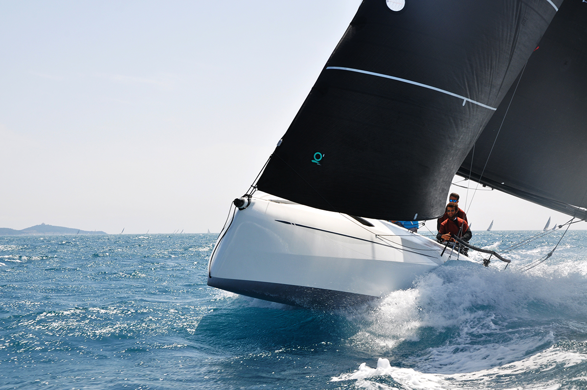 A person sailing a modern white sailboat with large black sails on the open blue ocean, leaning into the wind as waves splash against the hull under a clear sky.