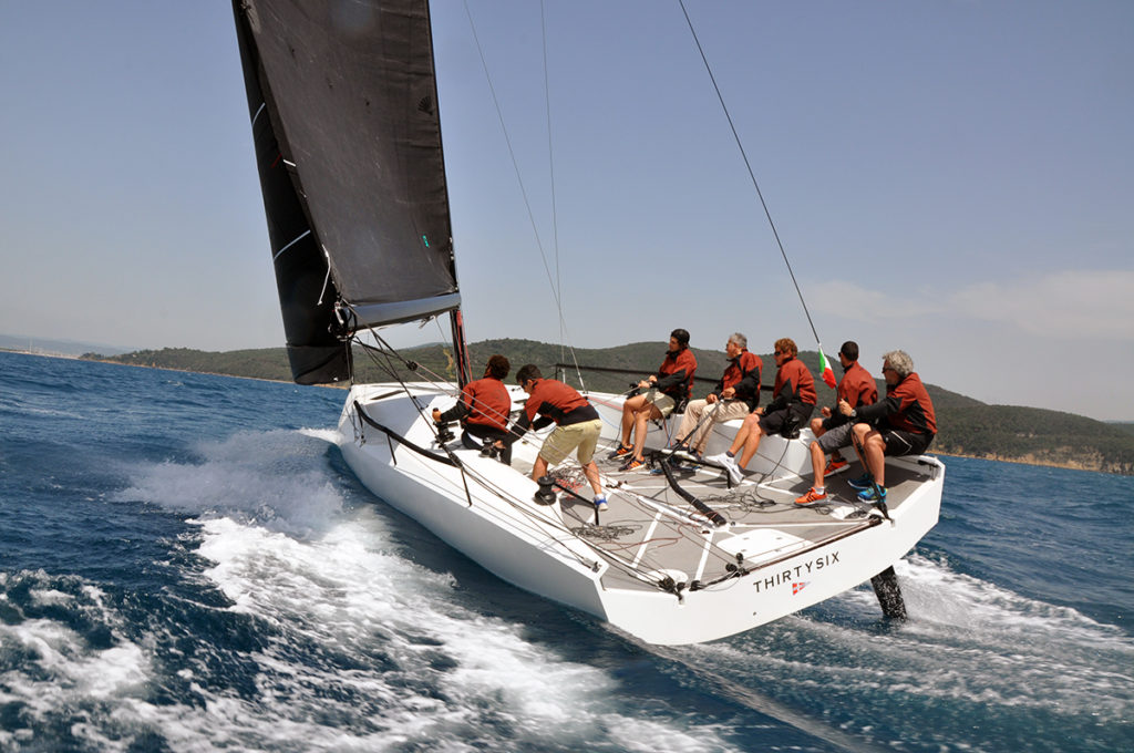 A team of eight people wearing matching red shirts sail a white racing yacht named 'THIRTYSIX' on blue water, leaning to one side as they speed through waves under a clear sky.