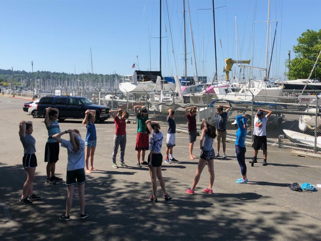 A group of people stand in a circle with their hands on their heads, exercising outdoors near a marina filled with sailboats on a sunny day. A fence and parked vehicles are visible in the background.
