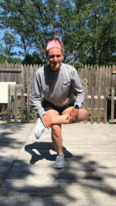 A person wearing a gray long-sleeve shirt, shorts, and sneakers stretches outdoors on a wooden deck, balancing on one leg with the other leg bent, and holding their ankle. Trees and a picket fence are in the background.