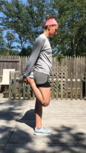 A person in athletic wear stands outdoors on a wooden deck, holding one foot behind them to stretch their leg. They wear a pink headband, gray long-sleeve shirt, black shorts, and sneakers. Trees and a wooden fence are in the background.