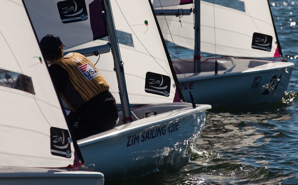Two small sailboats with “ZIM SAILING” branding are racing on the water. A person wearing a yellow USA vest is in the left boat, with sails catching the sunlight. The water is rippling around the boats.