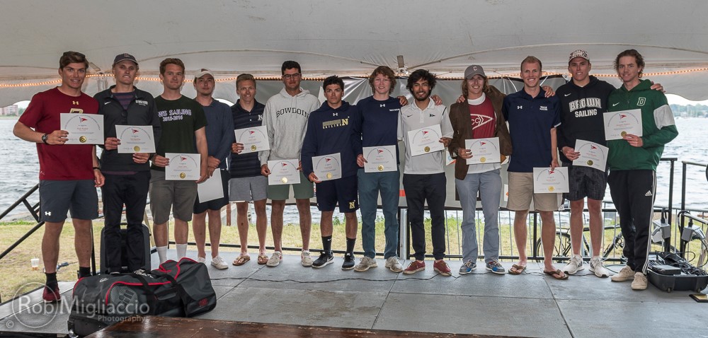 A group of 14 people stand in a line under a tent by the water, holding certificates and smiling at the camera. Most are wearing athletic or sailing-themed clothing. A duffel bag sits on the ground in front of them.