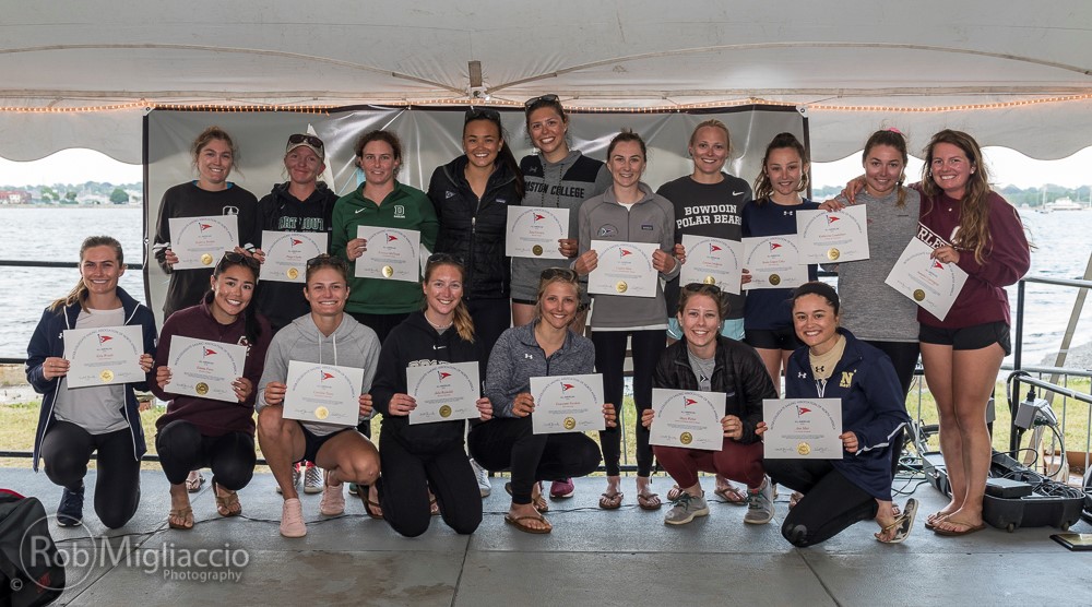 A group of women rowers pose under a tent by the water, smiling and holding certificates. Some are kneeling in front while others stand behind them. They are dressed in casual athletic clothing.