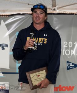 A young man in a navy St. Mary's Seahawks hoodie and cap holds a plaque while speaking into a microphone at an indoor award event. A sailboat graphic and sponsor logos are visible in the background.