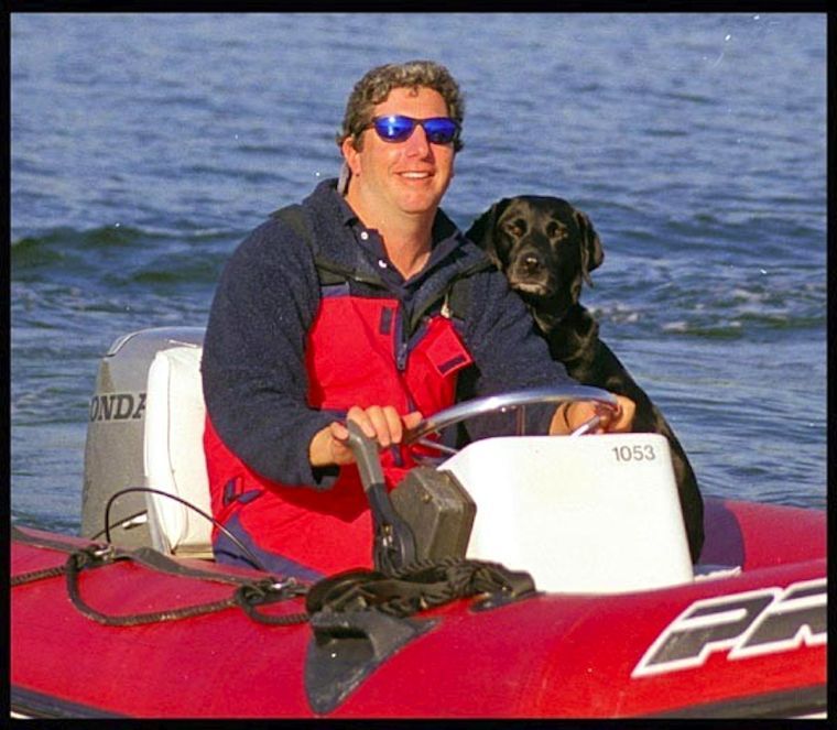 A smiling man wearing sunglasses and a red jacket drives a red inflatable boat on the water, with a black dog sitting beside him, both looking forward.