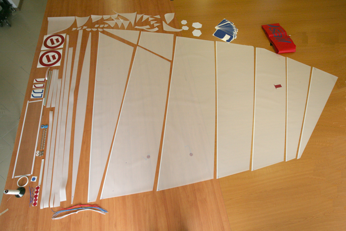 Overhead view of neatly arranged parts for assembling a sailboat sail, including white sail panels, various shapes of stickers, ropes, tape, and instruction booklets on a wooden table.