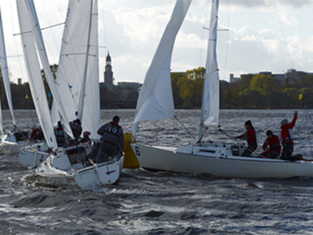 Four sailboats with crews race close together on a windy body of water, rounding a yellow buoy marked "6," with a cityscape and trees visible in the background.