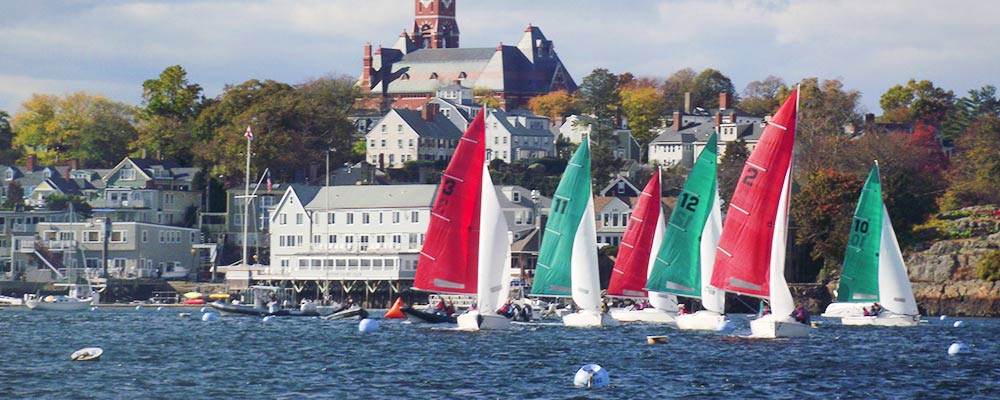 Colorful sailboats with red and green sails race on the water near a coastal town, with white buildings and a large brick church visible in the background among trees with autumn foliage.