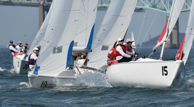 Several teams of sailors wearing life jackets compete in a sailboat race on choppy water, with white sails and a bridge visible in the background.