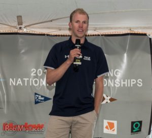 A man in a navy blue "Yale Sailing" polo shirt holds a microphone and speaks in front of a banner reading "2021 National Championships." Logos and the text "Rob Migliaccio Photography" are visible on the banner.